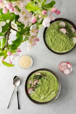 Top view of a table with two plates of creamy spinach broccoli soup, soup decorated with microgreens and sesame seeds with radishes, a bouquet of flowering apple tree branchesの写真素材