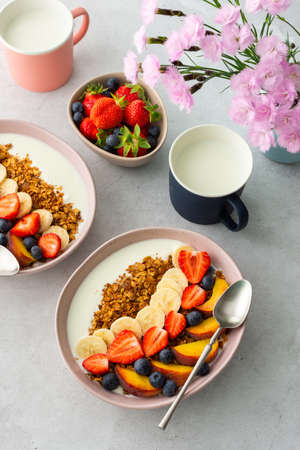 Delicious healthy breakfast, granola with yogurt and sweet fruits, banana and strawberries, blueberries and peaches, milk in a mug and flowers in a vase, top viewの写真素材