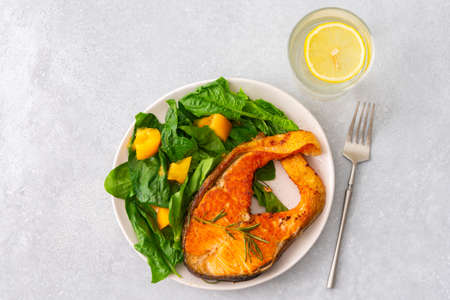 Fried salmon steak with spinach and yellow tomatoes in a plate on a gray background, a glass of water with lemon, a hearty lunch with healthy red fishの写真素材