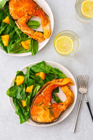 Fried salmon steak with spinach and yellow tomatoes in a plate on a gray background, a glass of water with lemon, a hearty lunch with healthy red fishの写真素材