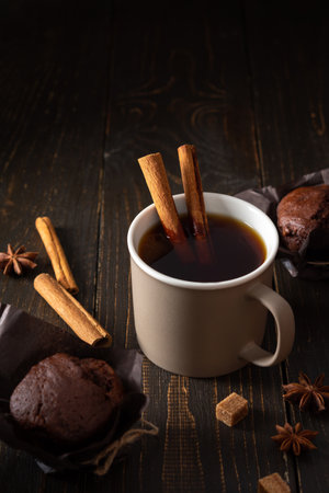 Cream ceramic cup with black tea and brown sugar, cinnamon and star anise on a dark wooden background, delicious chocolate muffins for teaの写真素材