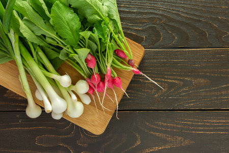 Freshly picked young radishes with green tops and green onions on a board on a brown wooden background, organic products from the vegetable gardenの写真素材