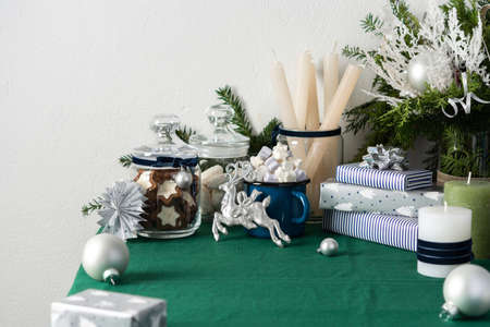 Festive decorated Christmas or New Year's table, thuja and decorative spruce branches, candles and Christmas balls, cookies in a jar and a blue iron mug with marshmallows, gift boxes and silver deerの写真素材