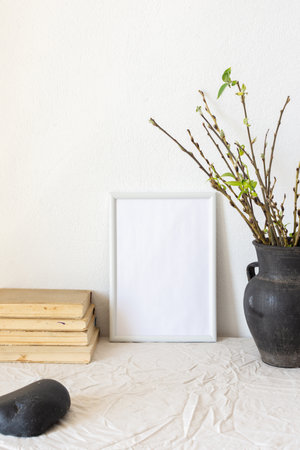 Home decor mock-up, blank picture frame near white painted concrete wall , black earthenware jug with pussy willow branches and blossoming green leaves, old booksの写真素材