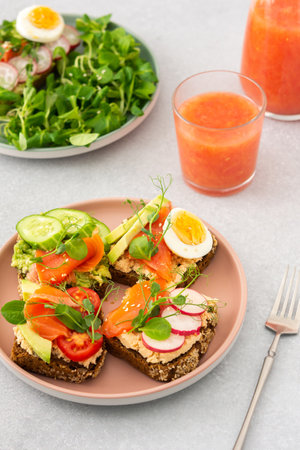 Healthy tasty breakfast, sandwiches with avocado and salmon, cucumbers and tomatoes, radishes and microgreens of peas with boiled egg and root salad, grapefruit smoothie in a glassの写真素材