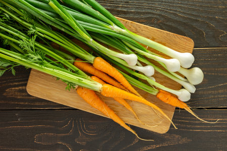 Freshly picked young carrots with green tops and green onions on a board on a brown wooden background, organic products from the vegetable gardenの写真素材