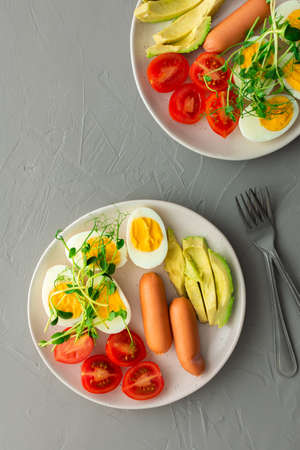 Delicious breakfast, boiled eggs with sausages, tomatoes and avocado, microgreens of peas and sunflowers, plates with food on a gray background ,top viewの写真素材
