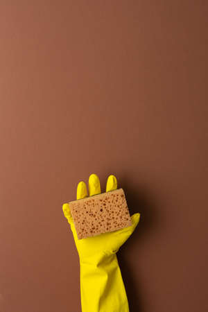 A hand in a yellow protective rubber glove holds a washing sponge on a brown backgroundの写真素材