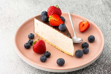 A piece of cheesecake covered with chocolate with blueberries and strawberries close-up on a pink plate with a dessert spoonの写真素材