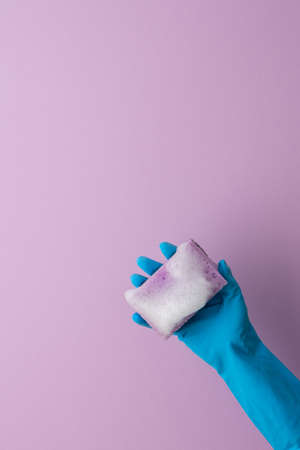 A hand in a blue protective rubber glove holds a washing sponge with foam on a purple backgroundの写真素材