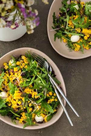 Salad with young boiled corn and mozzarella cheese, arugula and microgreen radish, fresh summer salad and flowers in a vase, top viewの写真素材