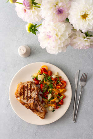 Delicious hearty dinner in plates on the table and a bouquet of beautiful autumn flowers, fried pork entrecote on the bone with stewed vegetables and fresh salad with mozzarellaの写真素材