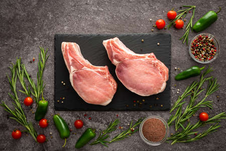 Black rectangular slate board with pork entrecote on the bone on gray background surrounded by fresh cherry tomatoes, jalapeno peppers, spices and rosemaryの写真素材