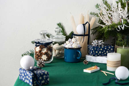 Festive decorated Christmas or New Year's table, thuja and decorative spruce branches, candles and white balls, cookies in a jar and a blue iron mug with marshmallows, gift boxes and fir conesの写真素材