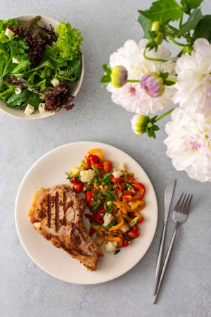 Delicious hearty dinner in plates on the table and a bouquet of beautiful autumn flowers, fried pork entrecote on the bone with stewed vegetables and fresh salad with mozzarellaの写真素材