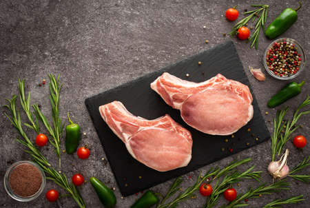 Black rectangular slate board with pork entrecote on the bone on gray background surrounded by fresh cherry tomatoes, jalapeno peppers, spices and rosemaryの写真素材