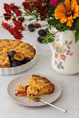 Homemade plum cake with almond flakes, autumn flowers in a vase, a piece of cake on a plateの写真素材