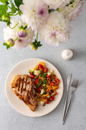 Delicious hearty dinner in plates on the table and a bouquet of beautiful autumn flowers, fried pork entrecote on the bone with stewed vegetablesの写真素材