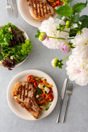 Delicious hearty dinner in plates on the table and a bouquet of beautiful autumn flowers, fried pork entrecote on the bone with stewed vegetables and fresh salad with mozzarellaの写真素材