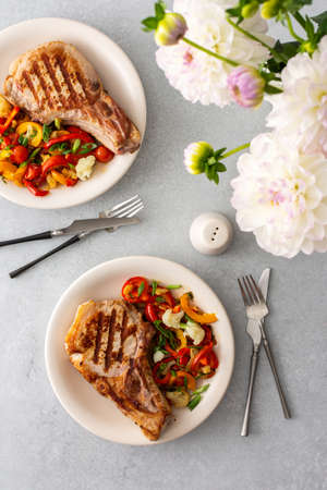 Delicious hearty dinner in plates on the table and a bouquet of beautiful autumn flowers, fried pork entrecote on the bone with stewed vegetablesの写真素材