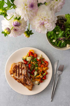 Delicious hearty dinner in plates on the table and a bouquet of beautiful autumn flowers, fried pork entrecote on the bone with stewed vegetables and fresh salad with mozzarellaの写真素材
