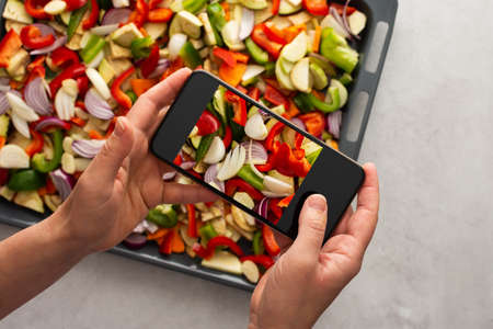 An adult woman takes pictures of stewed vegetables on the phone for social networks or a culinary blog, top view, home cooking hobby, healthy lifestyleの写真素材