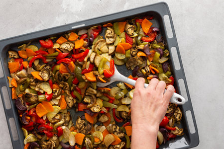 Woman stirring stewed vegetables with a silicone spatula, assorted autumn cut vegetables on a baking sheet, top viewの写真素材