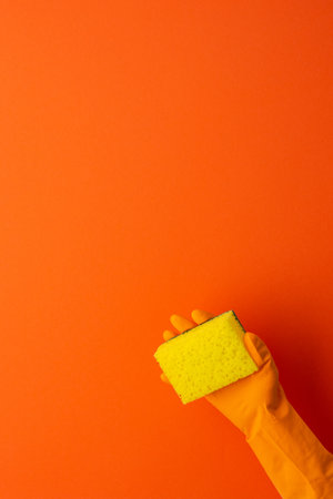 A hand in a protective rubber glove holds a yellow washing sponge on a orange backgroundの写真素材