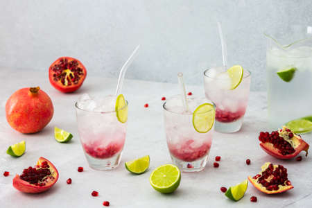 Refreshing pomegranate drink in glasses with pomegranate seeds and ice, lime and glass straws, jug of cold water with citrus, cut pomegranates with seedsの写真素材