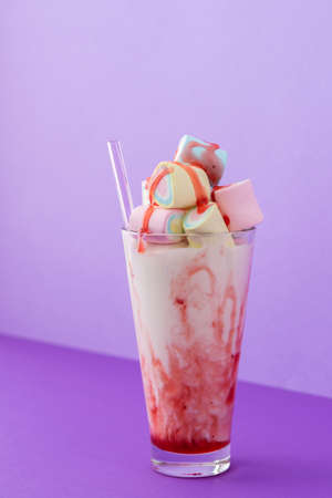 Glass cup with milkshake and fruit syrup, multicolored marshmallows and glass straws on bright purple background, holiday or birthday conceptの写真素材