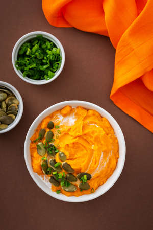 Pumpkin puree soup of butternut squash in bowl with green onions and pumpkin seeds, top view on plate with food on brown backgroundの写真素材