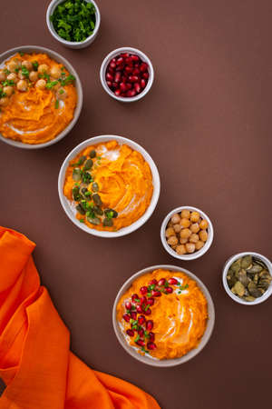 Pumpkin puree soup of butternut squash in bowls with chickpeas and green onions, pumpkin and pomegranate seeds, top view on plates with food on brown backgroundの写真素材