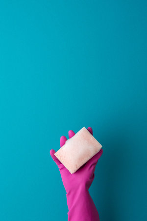 A hand in a purple protective rubber glove holds a pink washing sponge with foam on a blue backgroundの写真素材