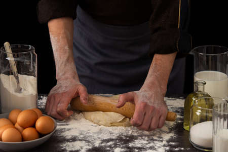 A man baker kneads dough on the table, milk in a jug and flour, chicken eggs in a plate and sugar and butter, male hobby for cookingの写真素材