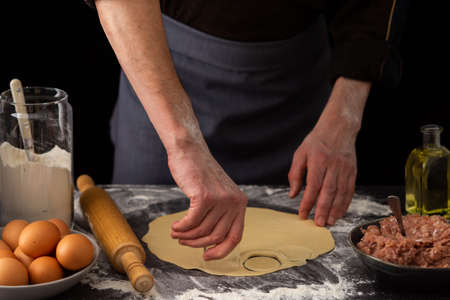 A man cuts out a mold for a russian traditional dish dumplings, ingredients for the dough and minced meat on the tableの写真素材