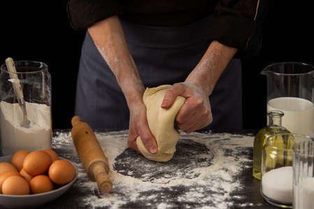 A man baker kneads dough on the table, milk in a jug and flour, chicken eggs in a plate and sugar and butter, male hobby for cookingの写真素材