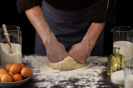 A man baker kneads dough on the table, milk in a jug and flour, chicken eggs in a plate and sugar and butter, male hobby for cookingの写真素材