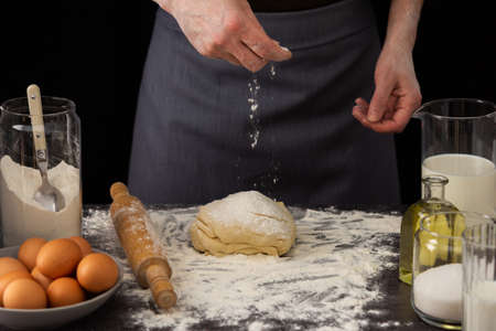 A man baker kneads dough on the table, milk in a jug and flour, chicken eggs in a plate and sugar and butter, male hobby for cookingの写真素材