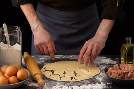 A man cuts out a mold for a russian traditional dish dumplings, ingredients for the dough and minced meat on the tableの写真素材
