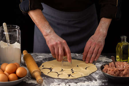 A man cuts out a mold for a russian traditional dish dumplings, ingredients for the dough and minced meat on the tableの写真素材