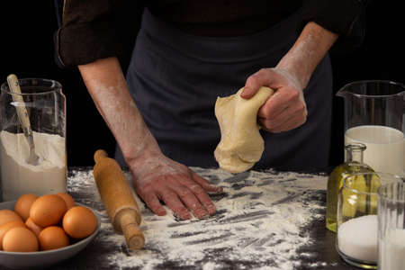 A man baker kneads dough on the table, milk in a jug and flour, chicken eggs in a plate and sugar and butter, male hobby for cookingの写真素材