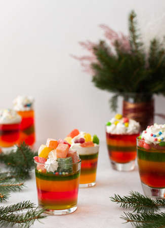 Glasses with multi-colored jelly, decorated with cream and dragees, meringues and marshmallows, Turkish delight and marmalade, drinks among the fir branches on the holiday tableの写真素材