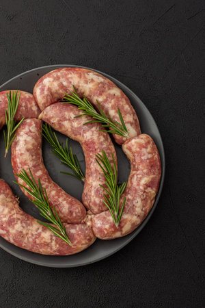 Raw pork sausages with rosemary in a plate close-up on a dark background