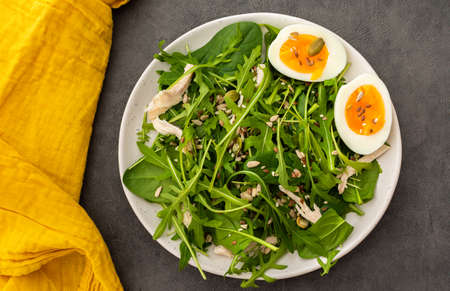 Delicious light salad with arugula and spinach, boiled eggs and chicken breast sprinkled with various healthy seeds in a plate on a gray background, top viewの写真素材