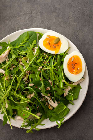 Delicious light salad with arugula and spinach, boiled eggs and chicken breast sprinkled with various healthy seeds in a plate on a gray background, top viewの写真素材