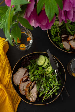 Hearty delicious lunch, fresh salad with cucumbers and microgreens of peas and with stewed pork, top view of the dark table with food and a vase of flowersの写真素材