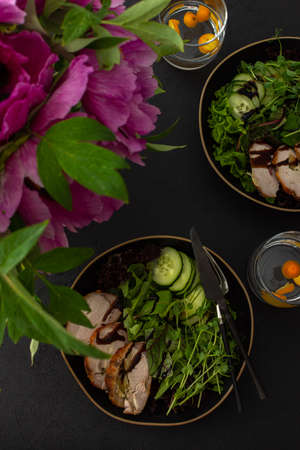 Hearty delicious lunch, fresh salad with cucumbers and microgreens of peas and with stewed pork, top view of the dark table with food and a vase of flowersの写真素材