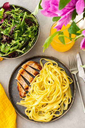 Hearty delicious lunch, pasta with fried chicken breast and fresh salad and orange juice, top view of the table with food and a vase of flowersの写真素材