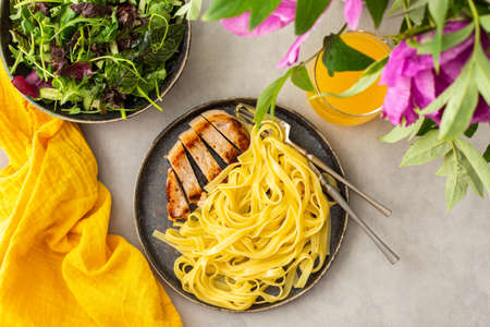Hearty delicious lunch, pasta with fried chicken breast and fresh salad and orange juice, top view of the table with food and a vase of flowersの写真素材