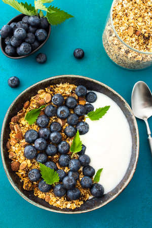 Homemade granola with yogurt, blueberries and mint leaves in a bowl on a turquoise background, delicious healthy breakfastの写真素材